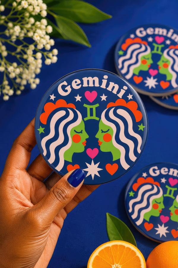 A close-up photograph of a round, glossy ceramic coaster featuring a bold retro pop art design. The background is a deep royal blue. In the center are two symmetrical profile faces of twins looking at each other, featuring bright green skin, closed eyes, and red cheeks. They have wavy white and pale pink hair with red accents. The word Gemini is arched at the top in a retro white font. The design is decorated with a green Gemini symbol, pink and red hearts, and white starbursts.
