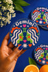 A close-up photograph of a round, glossy ceramic coaster featuring a bold retro pop art design. The background is a deep royal blue. In the center are two symmetrical profile faces of twins looking at each other, featuring bright green skin, closed eyes, and red cheeks. They have wavy white and pale pink hair with red accents. The word Gemini is arched at the top in a retro white font. The design is decorated with a green Gemini symbol, pink and red hearts, and white starbursts.