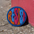 Round embroidered iron-on patch featuring "RESIST" text in bold flame design. Blue border surrounds navy background with orange, red, and blue flame-style lettering. 3x3 inch political activist patch with premium twill fabric construction, perfect for jackets, backpacks, and clothing customization. Anti-establishment protest accessory for resistance movements and political activism demonstrations.
