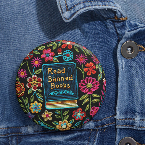 Round pinback button featuring a teal book with "Read Banned Books" in gold lettering on black background, surrounded by vibrant embroidery-style flowers in pink, orange, and blue. Literary protest pin with glossy finish and safety pin backing, available in 2" and 3" sizes for bookish fashion statements.