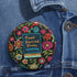 Round pinback button featuring a teal book with "Read Banned Books" in gold lettering on black background, surrounded by vibrant embroidery-style flowers in pink, orange, and blue. Literary protest pin with glossy finish and safety pin backing, available in 2" and 3" sizes for bookish fashion statements.