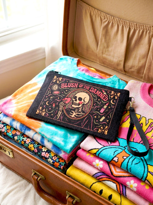 A rectangular gothic makeup bag made of black polyester with a visible woven linen-like crosshatch texture. The central illustration features a smiling human skull with flowing hair applying a pink heart-shaped blush to its cheek. Above the skull, the text BLUSH OF THE DAMNED appears. The design is framed by illustrations of lipstick tubes, mascara wands, bows. The bag includes a black zipper along the top edge and a removable black nylon wrist strap attached to the metal zipper pull.