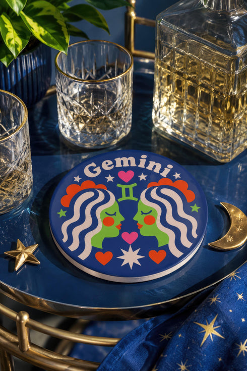 A close-up photograph of a round, glossy ceramic coaster featuring a bold retro pop art design. The background is a deep royal blue. In the center are two symmetrical profile faces of twins looking at each other, featuring bright green skin, closed eyes, and red cheeks. They have wavy white and pale pink hair with red accents. The word Gemini is arched at the top in a retro white font. The design is decorated with a green Gemini symbol, pink and red hearts, and white starbursts.