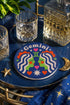 A close-up photograph of a round, glossy ceramic coaster featuring a bold retro pop art design. The background is a deep royal blue. In the center are two symmetrical profile faces of twins looking at each other, featuring bright green skin, closed eyes, and red cheeks. They have wavy white and pale pink hair with red accents. The word Gemini is arched at the top in a retro white font. The design is decorated with a green Gemini symbol, pink and red hearts, and white starbursts.
