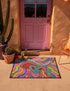 A rectangular doormat featuring a vibrant 2D flat print designed to look like textured yarn embroidery. The central graphic showcases a bright green alien with large black eyes, wearing a pink neck bandana and a highly decorated red, pink, and blue sombrero cowboy hat. The alien has one green hand raised in a friendly wave. The background is a psychedelic pattern of wavy, flowing rainbow stripes in yellow, orange, red, pink, blue, and green. The mat has a sleek black outer edge trim.