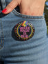 A round metal pinback button badge featuring a black background with the words QUEER MAGIC IS REAL in capital pink retro serif typography. A bright rainbow colored lightning bolt strikes from the top center, surrounded by concentric rainbow rings in red, orange, yellow, green, blue, and purple. The design is accented with small pink four-point stars and red crescent moons. The artwork features a realistic 2-D faux embroidery texture. The back has a silver metal safety pin clasp fastening.