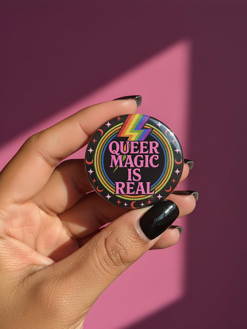 A round metal pinback button badge featuring a black background with the words QUEER MAGIC IS REAL in capital pink retro serif typography. A bright rainbow colored lightning bolt strikes from the top center, surrounded by concentric rainbow rings in red, orange, yellow, green, blue, and purple. The design is accented with small pink four-point stars and red crescent moons. The artwork features a realistic 2-D faux embroidery texture. The back has a silver metal safety pin clasp fastening.