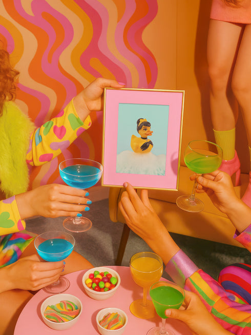 A chic rubber duck styled like Audrey Hepburn’s Holly Golightly, wearing a high bun, yellow bow, black gloves, and a sparkly choker. The duck floats in a pile of foam and is framed in white on a bubbly bathroom shelf with a red tile backdrop and soap dispenser.