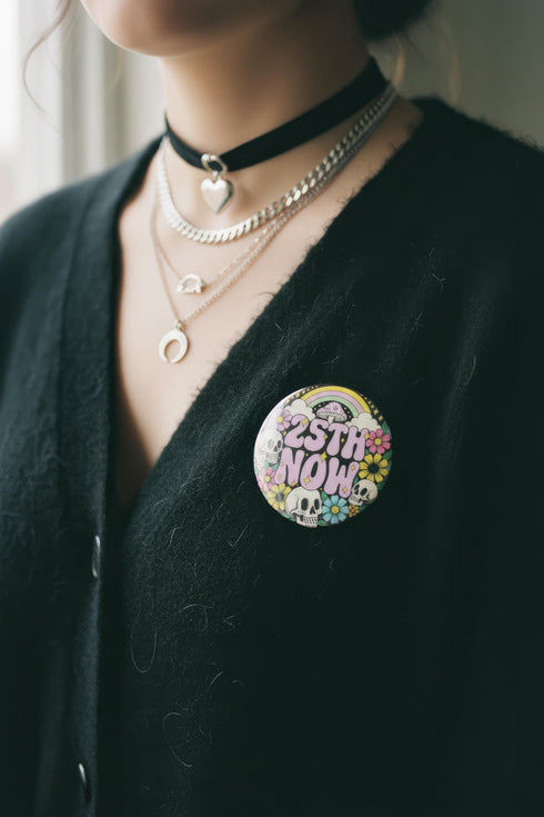 A round pinback button featuring a solid black background covered in a retro pastel colored design. The center reads 25TH NOW in a groovy bubbly font using pale pink and mint green referencing the 25th amendment. Surrounding the text is a maximalist cluster of cute but edgy motifs including smiling white skulls, pastel rainbows, pale yellow crescent moons, daisies, and a pink and white spotted mushroom. The button has a glossy laminate finish and a secure metal safety pin backing.