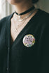 A round pinback button featuring a solid black background covered in a retro pastel colored design. The center reads 25TH NOW in a groovy bubbly font using pale pink and mint green referencing the 25th amendment. Surrounding the text is a maximalist cluster of cute but edgy motifs including smiling white skulls, pastel rainbows, pale yellow crescent moons, daisies, and a pink and white spotted mushroom. The button has a glossy laminate finish and a secure metal safety pin backing.