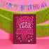 A birthday card with the text "Another Year Stranger" in a vibrant pink and purple color scheme, placed in front of a pink background with a "Happy Birthday" banner and green tinsel.
