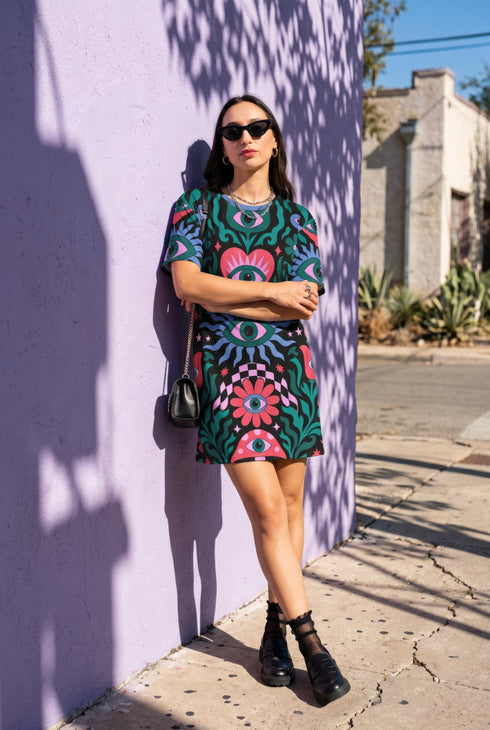 A short-sleeve, above-the-knee t-shirt dress with a true black background. The front features a vibrant, vertically stacked symmetrical graphic design. From top to bottom, the design includes a pink heart containing a green and blue eye, a large blue sunburst with a central eye, a red daisy flower with an eye flanked by pink and black checkered arches, and a pink mushroom cap with an eye. Swirling emerald green foliage, pink stars, and purple stars fill the negative space around the motifs.