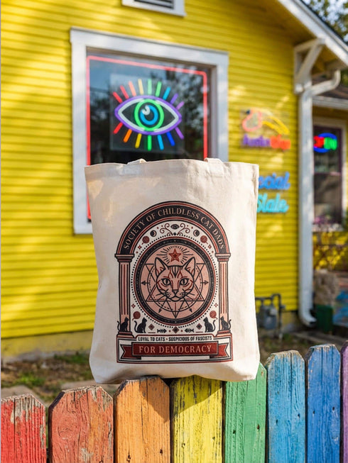 A beige canvas tote bag featuring a vintage-style circular crest design in rust orange and black. The text reads "Society of Childless Cat Dads" in an arch over a central illustration of a tabby cat face inside a geometric star and hexagon symbol. Below the cat, the text reads "Loyal to Cats, Suspicious of Fascists." A banner at the bottom reads "For Democracy." The design features occult and whimsigoth elements like crescent moons, columns, and fish bones.