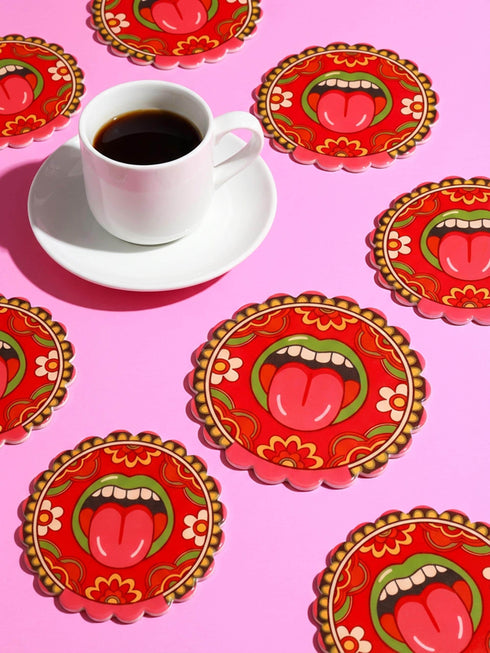 Round ceramic coaster with scalloped edges featuring a vibrant pop art design of an open mouth with green lips and a red tongue sticking out. The background is a bright red with psychedelic floral swirls in yellow orange and green. The surface is glossy glazed ceramic. The back is made of cork to prevent slipping. This maximalist decor piece fits retro 70s funky and weirdcore aesthetics. It is a bold statement piece for coffee tables. The design is colorful and eclectic dopamine decor.