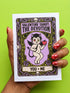 A vertical folded greeting card featuring a pastel goth tarot design titled The Devotion. The central illustration shows a pale devil imp with black wings and horns, holding a red heart in the left hand and a black dagger in the right. The imp has one star eye and one heart eye. The background is lavender purple. A black border frames the scene with teal vines, skulls, gold crescent moons, and roses. The bottom banner reads YOU + ME. The finish is shimmering pearlescent.