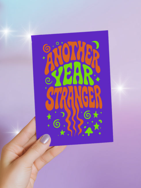 A hand holding a purple card with the text "Another Year Stranger" written on it.