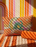 Rectangular 20x12 inch lumbar pillow cover in bright orange featuring printed faux embroidery of a green frog wearing a pink cowgirl hat and pink cowboy boots, lounging on a golden yellow crescent moon shaped like a peeled banana, surrounded by cream and gold sparkle stars and retro wavy stripes. Whimsical cowboy frog design printed on both sides with a hidden zipper, crafted from soft linen look polyester in a funky 70s retro style.