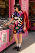 A short-sleeve, above-the-knee t-shirt dress with a relaxed, straight fit and a crew neckline. The dress features a vibrant, chaotic, all-over graphic print. The design includes sections of distorted, wavy black and cream checkerboard, mixed with bold, abstract shapes in bright purple, hot pink, and vivid orange. Scattered throughout the pattern are motifs of hot pink dripping slime, a black bat silhouette, a stylized green eye with a pink iris, and retro pink and purple daisy flowers.