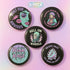 A set of five round 2.25-inch pinback buttons featuring original roller derby designs including a bruised face, saint on skates, flaming roller skate, skull in helmet, and bold text phrases like “Built for Bruises” and “Hell on Wheels.” Each button has a glossy black finish with vibrant pastel pink, purple, mint, and aqua artwork, mounted on metal tinplate with a safety pin back.