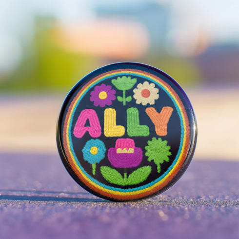 Vibrant ALLY pinback button featuring 70s-style embroidered flowers and lettering in pink, yellow, green, and blue against a black background, surrounded by a rainbow border. A colorful statement accessory combining punk activism and retro flower power aesthetics for backpacks, jackets, and protest wear.