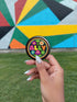 Vibrant ALLY pinback button featuring 70s-style embroidered flowers and lettering in pink, yellow, green, and blue against a black background, surrounded by a rainbow border. A colorful statement accessory combining punk activism and retro flower power aesthetics for backpacks, jackets, and protest wear.
