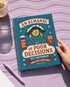 A spiral-bound hardcover planner featuring a retro-style teal cover with the text "An Almanac of Poor Decisions" in bold red and cream typography. The cover includes a grumpy cloud raining down lightning bolts, alongside stars, flowers, and vintage banners. The interior reveals crisp white pages with a 2026 to 2027 year-at-a-glance spread, plus clean, minimalist, completely undated monthly and weekly calendar grids with dedicated sections for writing daily to-do lists and random notes.
