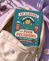 A spiral-bound hardcover planner featuring a retro-style teal cover with the text "An Almanac of Poor Decisions" in bold red and cream typography. The cover includes a grumpy cloud raining down lightning bolts, alongside stars, flowers, and vintage banners. The interior reveals crisp white pages with a 2026 to 2027 year-at-a-glance spread, plus clean, minimalist, completely undated monthly and weekly calendar grids with dedicated sections for writing daily to-do lists and random notes.