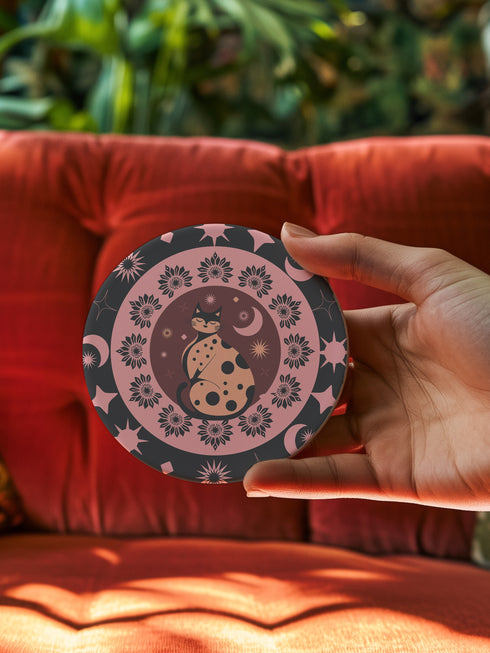Round ceramic coaster featuring a stylized black cat with polka dots surrounded by pink flowers, moons, and stars on a black background with pink accents