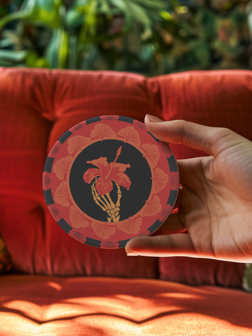 Round ceramic coaster featuring a skeleton hand holding a coral hibiscus flower, surrounded by art deco fan patterns in coral and black
