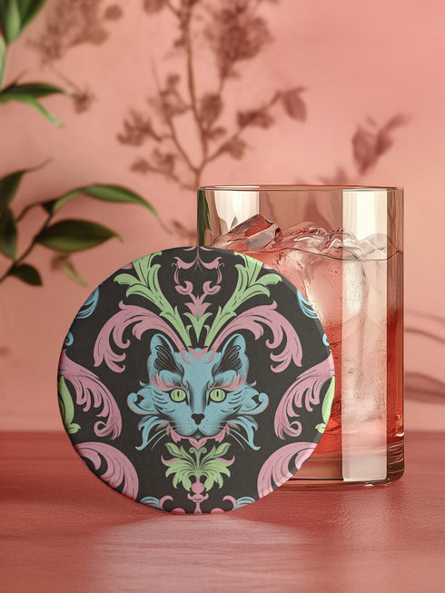 Circular ceramic coaster featuring a turquoise cat face with green eyes on black background, surrounded by pink scrollwork and green leaves in Victorian damask style.