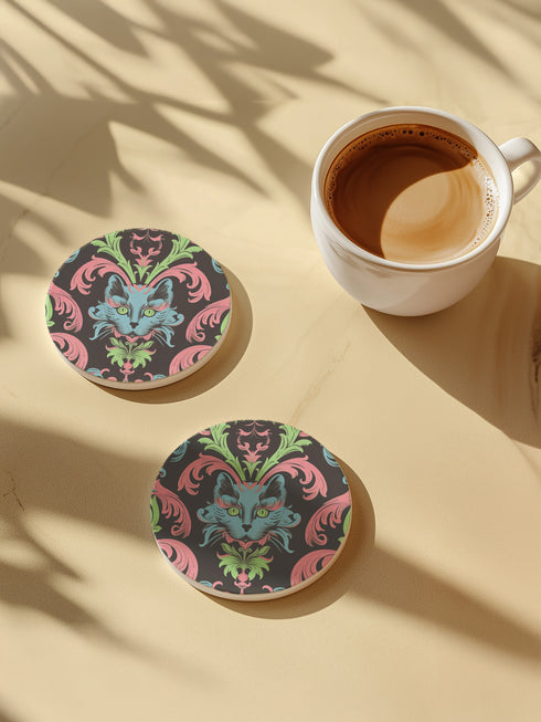 Circular ceramic coaster featuring a turquoise cat face with green eyes on black background, surrounded by pink scrollwork and green leaves in Victorian damask style.