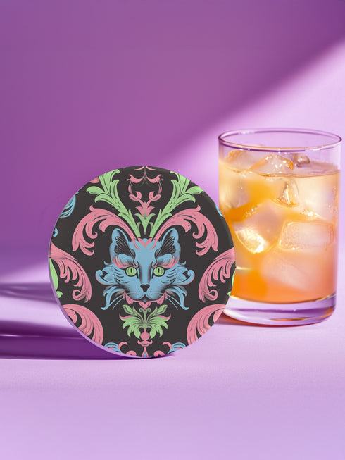 Circular ceramic coaster featuring a turquoise cat face with green eyes on black background, surrounded by pink scrollwork and green leaves in Victorian damask style.