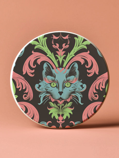 Circular ceramic coaster featuring a turquoise cat face with green eyes on black background, surrounded by pink scrollwork and green leaves in Victorian damask style.