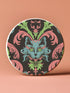 Circular ceramic coaster featuring a turquoise cat face with green eyes on black background, surrounded by pink scrollwork and green leaves in Victorian damask style.