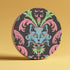 Circular ceramic coaster featuring a turquoise cat face with green eyes on black background, surrounded by pink scrollwork and green leaves in Victorian damask style.
