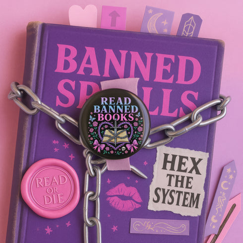 Glossy black pinback button that says “Read Banned Books” in purple and teal letters, featuring a chained book, roses, bows, and floral accents, styled on colorful books and pastel backgrounds.