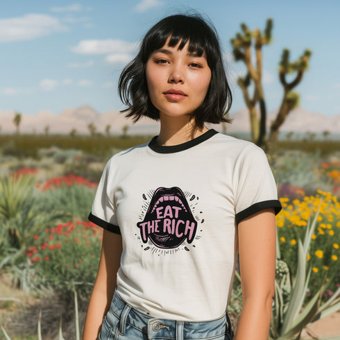 Off-white ringer t-shirt with black contrast trim on the collar and sleeve cuffs. The front features a bold, high-contrast graphic of an open mouth with the phrase "EAT THE RICH" in capital letters, styled in a hand-drawn font. The design is rendered in black and vibrant pink, with expressive line details surrounding the mouth, adding a dynamic, rebellious feel. The t-shirt has a vintage-inspired fit, made from soft cotton fabric with a slightly relaxed silhouette. The black trim enhances the retro aestheti