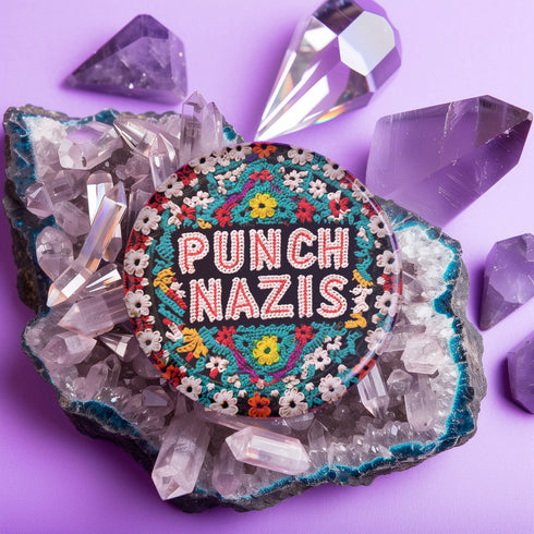 Round pinback metal button with an embroidered-style design on a black background. The center displays the phrase "PUNCH NAZIS" in bold, white stitched-textured lettering with red accents. Surrounding the text is an elaborate folk-inspired floral pattern, densely packed with embroidered-style flowers in shades of red, yellow, blue, pink, and white. The intricate design includes symmetrical lace-like stitching and decorative motifs, creating a rich, tapestry-like effect.