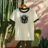 Unisex ringer t-shirt in natural cotton with black ribbed collar and sleeve cuffs. Front features bold black graphic: a cult-style goose surrounded by four candles, with text reading “Silly Goose Cult, Weekly Chaos, Snacks Provided.” Available in sizes S to 3XL. Retro-inspired fit, soft cotton, statement weirdcore design for alternative fashion lovers.