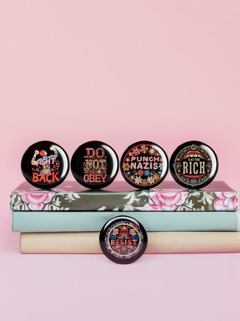 A set of five round, glossy black pinback buttons with embroidered folk art designs and bold activist slogans. Each pin features a different phrase such as "Eat the Rich" and "Resist," surrounded by colorful floral motifs.