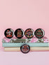 A set of five round, glossy black pinback buttons with embroidered folk art designs and bold activist slogans. Each pin features a different phrase such as "Eat the Rich" and "Resist," surrounded by colorful floral motifs.