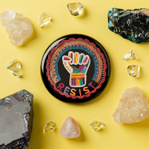 Round pinback metal button with an embroidered-style design on a black background. The center features a raised fist symbol stitched in a multicolored pattern, incorporating rainbow hues and folk-inspired textures. The fist is encircled by a vibrant, lace-like border in shades of blue, orange, and red, resembling intricate embroidery. Below the fist, the word "RESIST" is spelled out in bold, stitched-textured letters in red and orange. The button has a silver metal back with a secure pin closure.