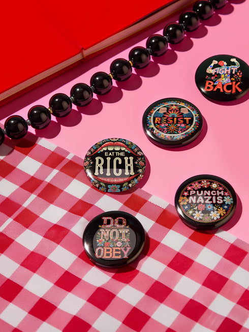 A set of five round, glossy black pinback buttons with embroidered folk art designs and bold activist slogans. Each pin features a different phrase such as "Eat the Rich" and "Resist," surrounded by colorful floral motifs.