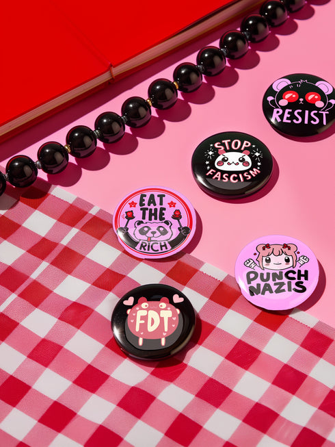 A set of five glossy round pinback buttons with pastel goth and kawaii punk designs, featuring slogans like "Eat The Rich" and "Stop Fascism" in bold pink and black. Each pin has a cute character with a rebellious message.