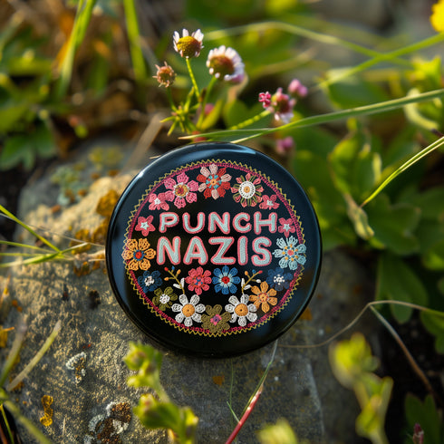 Round pinback metal button featuring an embroidered-style design on a black background. The center showcases bold, stitched-textured lettering in red, blue, and white that reads "PUNCH NAZIS." Surrounding the text is an intricate, folk-inspired floral pattern with multicolored flowers in shades of pink, orange, blue, and white, arranged symmetrically. A decorative zigzag border frames the edge of the button. The backside is silver metal with a secure pin closure.