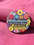 A round pinback button featuring a flat-printed design that mimics the appearance of highly detailed, 3D embroidery (trompe-l'œil). The background is solid black, with a floral border including red, yellow, sky blue, magenta, and orange flowers. In the center, the text "Billionaires Don't Exist in Nature" is written in a blue serif font. Below the text is a yellow rectangle with black borders containing a warning icon and the words "Warning: Invasive Species" in a bold, sans-serif black font.