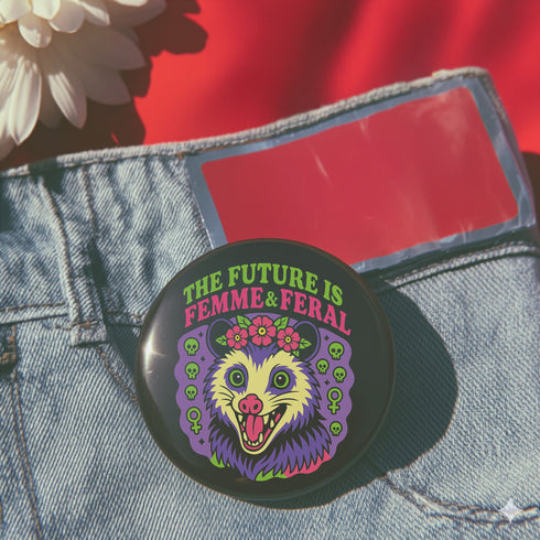 A round, black pinback button with a high-gloss finish features the text "THE FUTURE IS FEMME & FERAL" in a retro font of lime green and pastel pink. In the center, a screaming possum with wild eyes and a pink-lined mouth is illustrated in a pastel goth style with purple, yellow, and pink fur. The possum wears a pink flower crown. The illustration is framed by a circle of small skulls and pink Venus (female) symbols on a purple background. This is a piece of feminist possumcore flair.