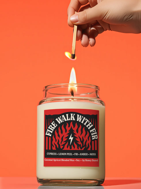 A 9-ounce hand-poured candle inspired by the TV show Twin Peaks, named "Fire Walk With Fir." The candle is made from a creamy, off-white coconut apricot blended wax, housed in a clear glass vessel with a classic, slightly tapered shape. A prominent matte-finish label on the front features the title in a stylized font over a red and black graphic with flame and fir tree motifs. The label also lists scent notes of cypress, lemon peel, fir, amber, and moss.