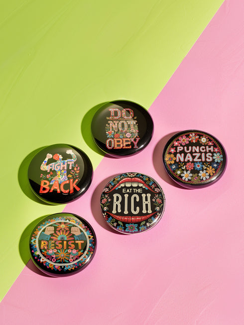 A set of five round, glossy black pinback buttons with embroidered folk art designs and bold activist slogans. Each pin features a different phrase such as "Eat the Rich" and "Resist," surrounded by colorful floral motifs.