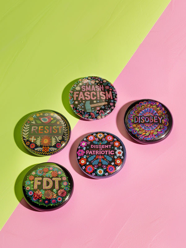 A set of five glossy black tinplate pinback buttons featuring intricate folk-art-inspired floral designs and bold activist messages such as "Smash Fascism," "Dissent is Patriotic," and "Resist."